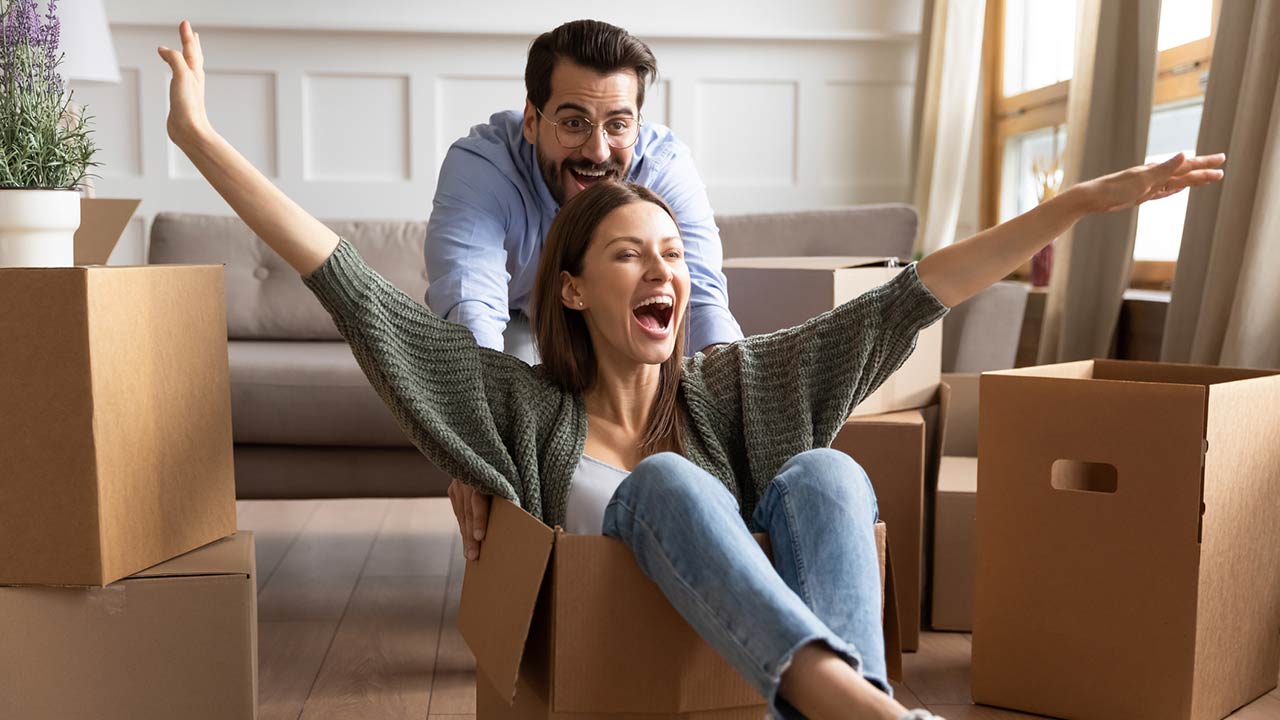 Happy family celebrating moving day, playful young couple having fun at home, funny activity, laughing husband pushing excited wife sitting in cardboard box