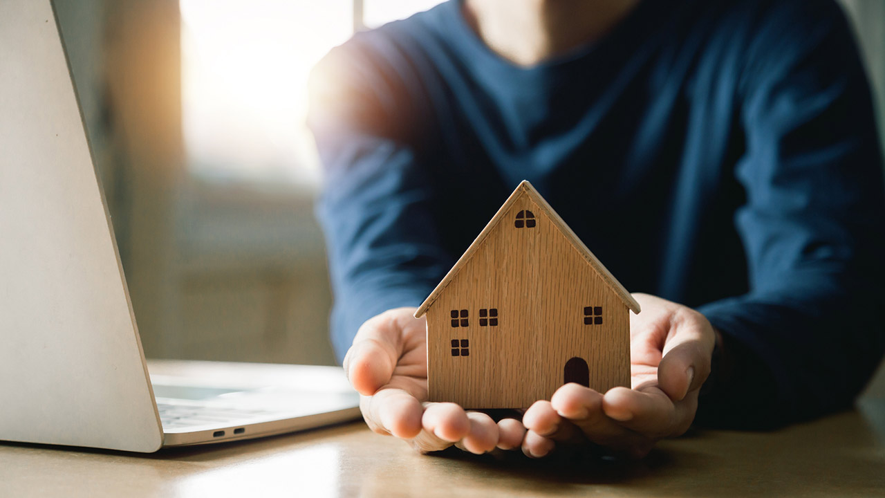 Hands holding a wooden model home with an open laptop